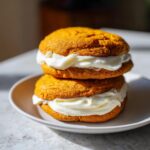 Two soft pumpkin cookie sandwiches filled with thick cream cheese frosting stacked on a white plate.