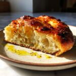 Close-up of a thick slice of golden-brown sourdough discard pizza dough, showing an airy crumb and olive oil.