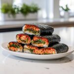 A stack of six pieces of spicy tuna kimbap, showing rice, greens, and red tuna filling wrapped in dark seaweed.
