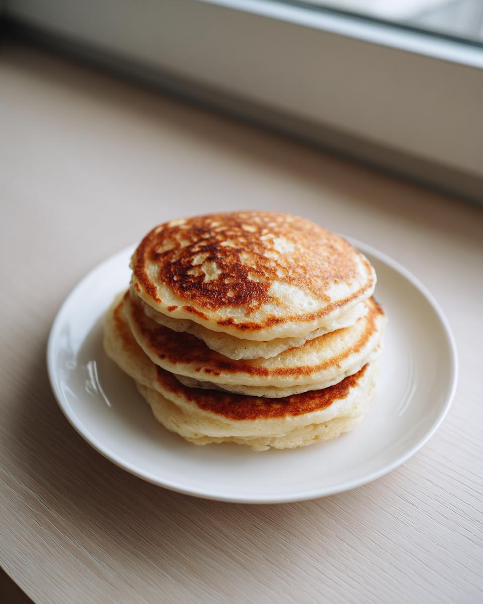 A stack of four perfectly golden brown banana oatmeal pancakes served on a white plate near a window.