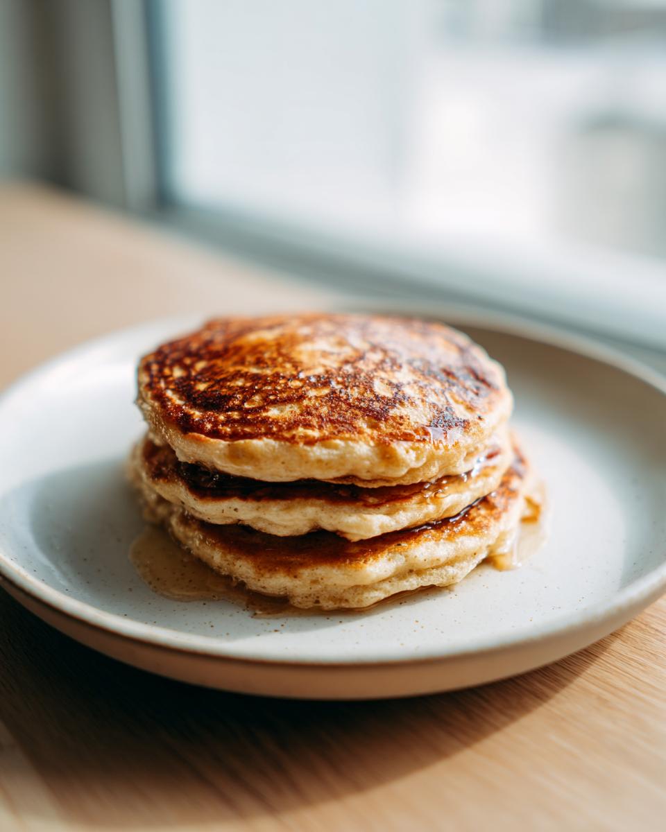 A close-up stack of three golden brown banana oatmeal pancakes drizzled with syrup on a light speckled plate.