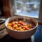 A steaming white bowl filled with rich, tomato-based pasta fagioli, set against a bright window.