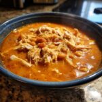 Close-up of a steaming blue bowl filled with rich, orange-red buffalo chicken chili topped with shredded chicken and beans.