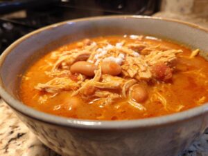 Close-up of a steaming bowl of rich orange buffalo chicken chili, topped with shredded chicken, white beans, and crumbled cheese.