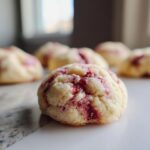 A close-up of a soft, pale cookie with swirls of bright red strawberry filling, one of several strawberry cheesecake cookies.