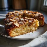 A moist slice of swedish apple cake, topped with toasted almonds and powdered sugar, served on a white plate.