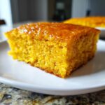 Close-up of a square slice of bright orange sweet potato cornbread, glistening with a honey glaze, served on a white plate.