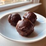 Three rich, chocolate-dipped Oreo balls resting on a small white plate near a window.