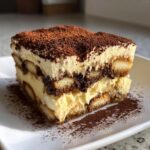 A rich, layered slice of tiramisu cheesecake topped generously with cocoa powder, served on a white plate.