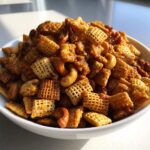 A white bowl filled with crunchy, glazed toffee chex mix recipe featuring cereal squares and whole cashews.