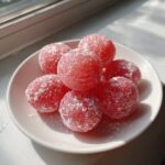 A small white plate holds several round, bright red watermelon mochi dusted lightly with powdered sugar.