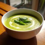 A bowl of vibrant green zucchini soup, swirled with cream and topped with fresh basil leaves.