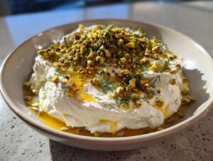 A bowl of creamy whipped feta dip topped generously with chopped pistachios, fresh dill, and a drizzle of golden honey.