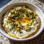 A creamy bowl of whipped feta dip topped generously with chopped pistachios, fresh dill, and drizzled honey.