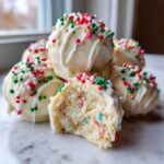 A pile of white chocolate coated sugar cookie truffles, one bitten to show the colorful interior, topped with red, green, and white sprinkles.