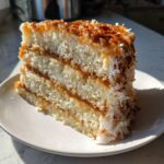 A tall slice of white German Chocolate Cake showing three layers of white cake separated by caramel-colored filling and covered in toasted coconut.
