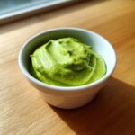 A small white bowl filled with creamy, bright green avocado sauce sitting on a wooden surface.