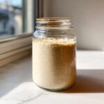 A bubbly, active sourdough starter filling a clear glass jar, sitting on a white marble surface near a window.