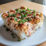 A square slice of amazing sushi bake served on a white plate, generously topped with creamy sauce, furikake, and green onions.