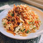 A vibrant mound of thai chicken salad featuring shredded cabbage, carrots, chicken, and topped with crushed peanuts and peanut dressing.