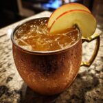 Close-up of a frosty copper mug filled with an apple cider moscow mule, garnished with fresh apple slices.