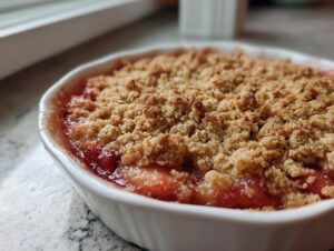 A close-up, angled view of a freshly baked rhubarb crisp with a golden, crumbly topping in a white ramekin.