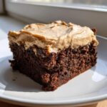 A close-up of a rich, dark chocolate banana bread brownies slice topped with thick, light brown frosting.