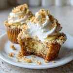 Close-up of a banana pudding cupcake cut in half showing creamy filling, topped with whipped cream and cookie crumbs.