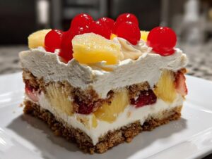 A close-up slice of banana split cake showing layers of graham cracker crust, cream filling, pineapple, strawberries, and topped with whipped cream and cherries.