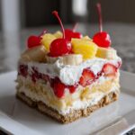 A perfect square slice of banana split cake showing layers of crust, cream, fruit, and whipped topping with cherries.
