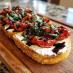 Two slices of toasted bread topped with creamy cheese, roasted tomatoes, fresh basil, and balsamic glaze, resembling a bruschetta dip serving.