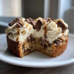 A close-up of a chocolate chip cookie cake recipe with a slice removed, topped with thick frosting and chopped candy bars.