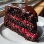 A decadent slice of chocolate raspberry cake showing dark layers, bright red raspberry filling, and glossy chocolate ganache on top.
