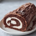 A close-up of a chocolate yule log cake showing the dark brown sponge rolled with white cream filling, frosted like bark.