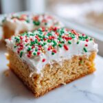 A close-up shot of a square slice of moist Christmas sugar cookie bars topped with thick white frosting and festive red, green, and gold sprinkles.