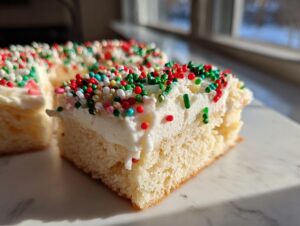 A close-up of a thick slice of Christmas sugar cookie bars topped with white frosting and festive red, green, and white sprinkles.