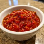 Close-up of a small white bowl filled with vibrant, chunky homemade tomato sauce, seasoned with herbs.
