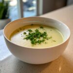 A close-up of a bowl of creamy celery soup, topped with fresh chopped parsley.