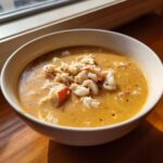 A close-up of a creamy, orange-hued bowl of crab soup topped generously with white crab meat and black pepper.