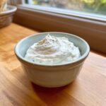 A small bowl filled with thick, creamy horseradish sauce, speckled with green herbs, sitting on a wooden counter.