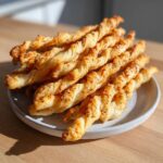 A close-up of a stack of golden, flaky, twisted cheese straws sprinkled with black pepper or seeds, served on a small plate.
