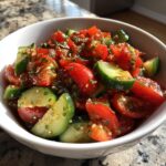 Close-up of a vibrant cucumber tomato salad with a dark, herby dressing in a white bowl.