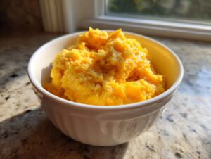 A close-up of a white bowl filled with creamy, bright orange cheese grits sitting on a granite countertop near a window.