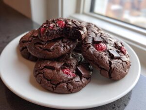 A stack of rich, dark chocolate cherry cookies with visible chocolate chunks and dried cherries, one cookie is broken open.