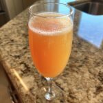 A close-up of a fizzy, orange bellini recipe cocktail served in a champagne flute on a granite countertop.