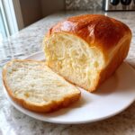 A loaf of freshly baked condensed milk bread, sliced open to show its soft, white interior, resting on a white plate.