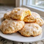 A stack of soft, glazed ricotta cookies on a white plate, with one cookie broken to show the fluffy interior.