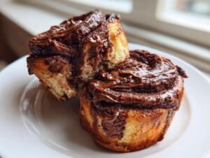 Two gooey chocolate cinnamon rolls stacked slightly on a white plate, showing rich chocolate filling.
