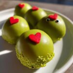 Five bright green Grinch Oreo balls decorated with small red hearts, resting on a light plate.