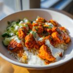 Close-up of a bowl filled with spicy harissa chicken over rice, topped with cooling yogurt sauce and chopped cucumbers.